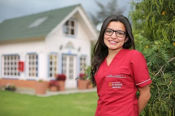 Joven sonriente con uniforme de medicina frente a una casa del albergue